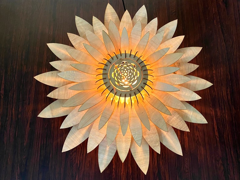 Op de afbeelding: Een houten lamp in de vorm van een zonnebloem met een gloeilamp in het midden. De lamp heeft een natuurlijke houten afwerking en wordt verlicht door een warm licht.