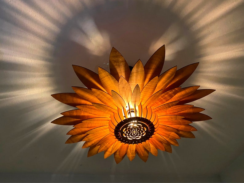 Puede incluir: Una l&aacute;mpara de techo de madera con forma de girasol con p&eacute;talos de color amarillo c&aacute;lido y un centro marr&oacute;n. La luz est&aacute; encendida, proyectando un brillo c&aacute;lido.