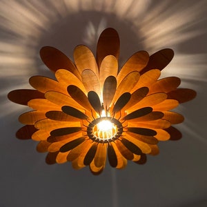 May include: A wooden ceiling light fixture shaped like a flower with multiple layers of petals. The light is on and casts a warm glow.