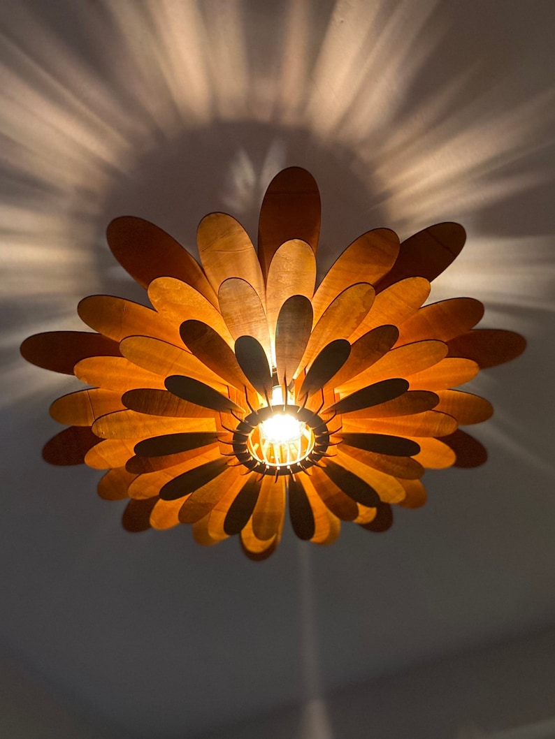 May include: A wooden ceiling light fixture shaped like a flower with multiple layers of petals. The light is on and casts a warm glow.