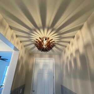 May include: A wooden ceiling light fixture with a unique design casts a shadow pattern on the ceiling. The light is in a hallway with a white door and gray wainscoting.