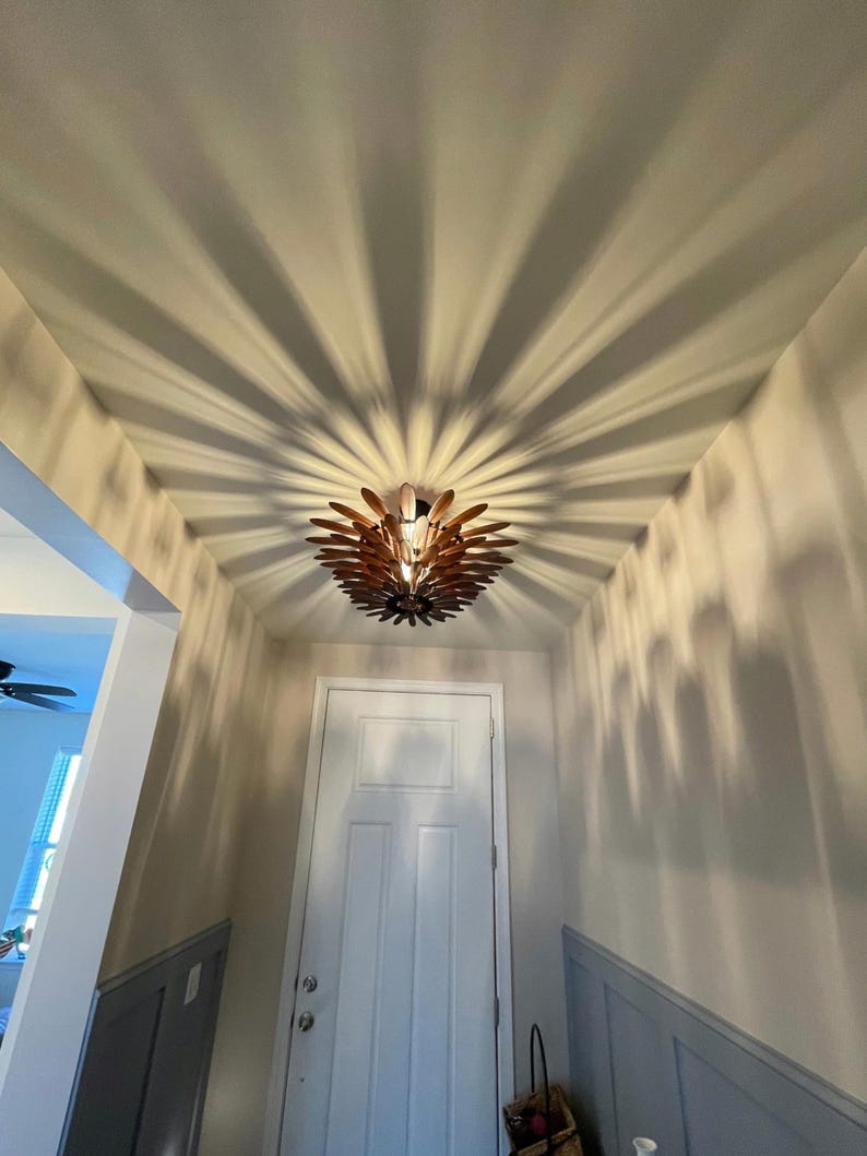 May include: A wooden ceiling light fixture with a unique design casts a shadow pattern on the ceiling. The light is in a hallway with a white door and gray wainscoting.