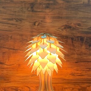 Op de afbeelding: Een houten tafellamp met een uniek, geometrisch ontwerp dat lijkt op een dennenappel. De lamp is aan en geeft een warm licht.