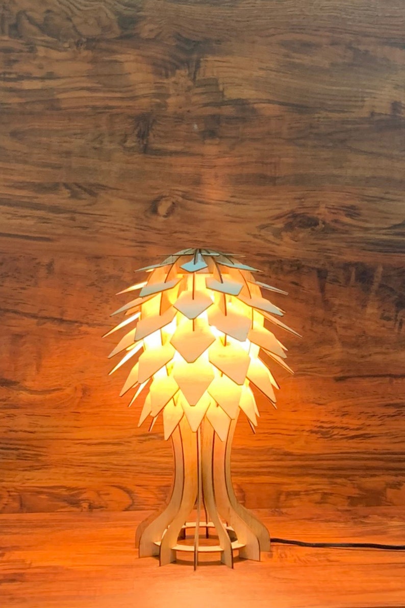 Op de afbeelding: Een houten tafellamp met een uniek, geometrisch ontwerp dat lijkt op een dennenappel. De lamp is aan en geeft een warm licht.