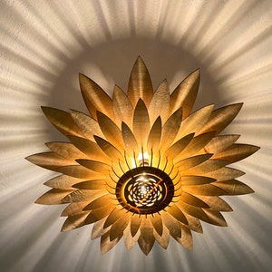 Op de afbeelding: Een gouden plafondlamp in de vorm van een zonnebloem met houten bloemblaadjes. Het licht is aan en geeft een warm gloed.