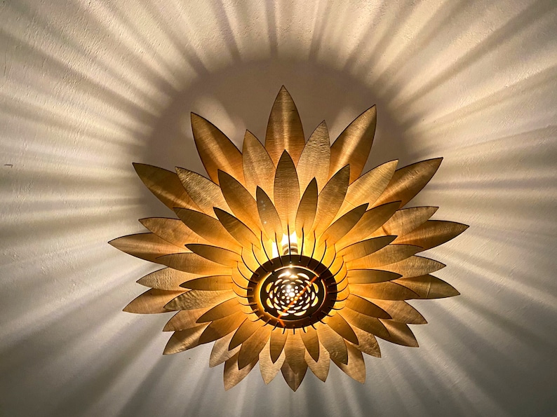 Op de afbeelding: Een gouden plafondlamp in de vorm van een zonnebloem met houten bloemblaadjes. Het licht is aan en geeft een warm gloed.