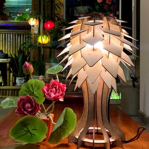 Peut inclure: Lampe de table en bois au design original, compos&eacute;e de panneaux en bois superpos&eacute;s cr&eacute;ant un abat-jour textur&eacute; et lumineux. La lampe est pos&eacute;e sur une table en bois, &agrave; c&ocirc;t&eacute; d'une plante en pot avec des fleurs rouges et des feuilles vertes.