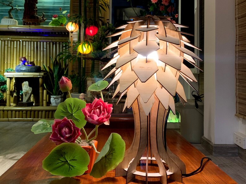 Peut inclure: Lampe de table en bois au design original, compos&eacute;e de panneaux en bois superpos&eacute;s cr&eacute;ant un abat-jour textur&eacute; et lumineux. La lampe est pos&eacute;e sur une table en bois, &agrave; c&ocirc;t&eacute; d'une plante en pot avec des fleurs rouges et des feuilles vertes.
