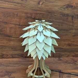 Op de afbeelding: Een houten tafellamp met een uniek, geometrisch ontwerp. De lampenkap is gemaakt van veel kleine, overlappende stukjes hout, waardoor een textuur en een organische uitstraling ontstaat. De voet van de lamp is ook van hout en heeft een eenvoudig, elegant ontwerp.