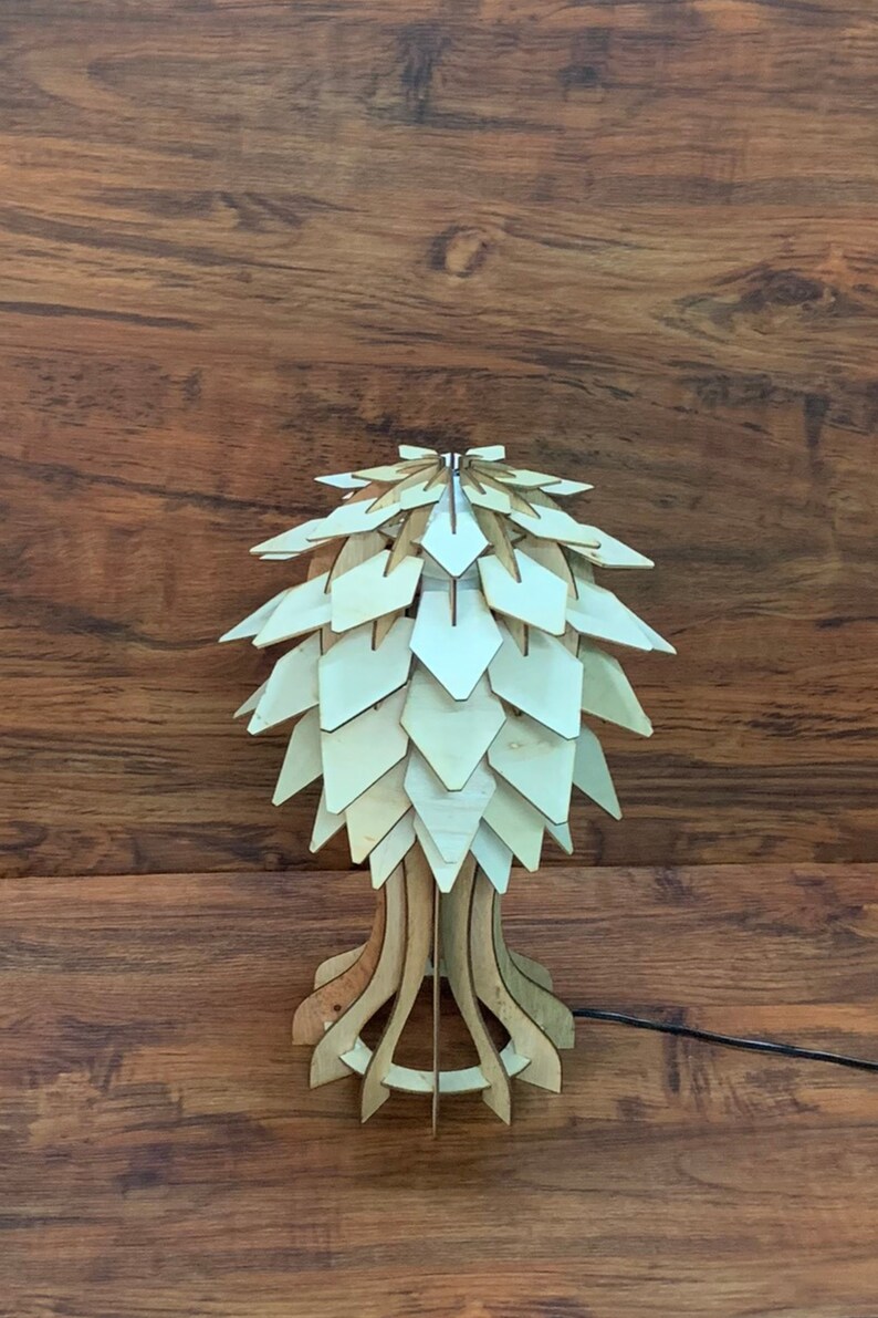 Op de afbeelding: Een houten tafellamp met een uniek, geometrisch ontwerp. De lampenkap is gemaakt van veel kleine, overlappende stukjes hout, waardoor een textuur en een organische uitstraling ontstaat. De voet van de lamp is ook van hout en heeft een eenvoudig, elegant ontwerp.