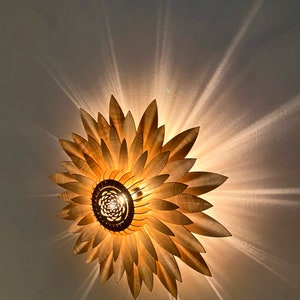 Op de afbeelding: Een houten plafondlamp in de vorm van een zonnebloem met een gloeilamp in het midden. De lamp geeft een warm licht op het witte plafond.