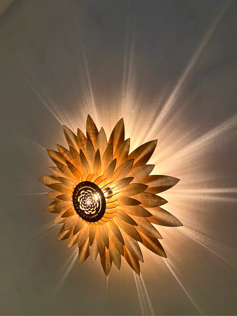 Op de afbeelding: Een houten plafondlamp in de vorm van een zonnebloem met een gloeilamp in het midden. De lamp geeft een warm licht op het witte plafond.