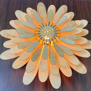 May include: A wooden ceiling light fixture shaped like a daisy with a light bulb in the center. The light fixture is made of many thin, overlapping pieces of wood that are painted a light brown color.