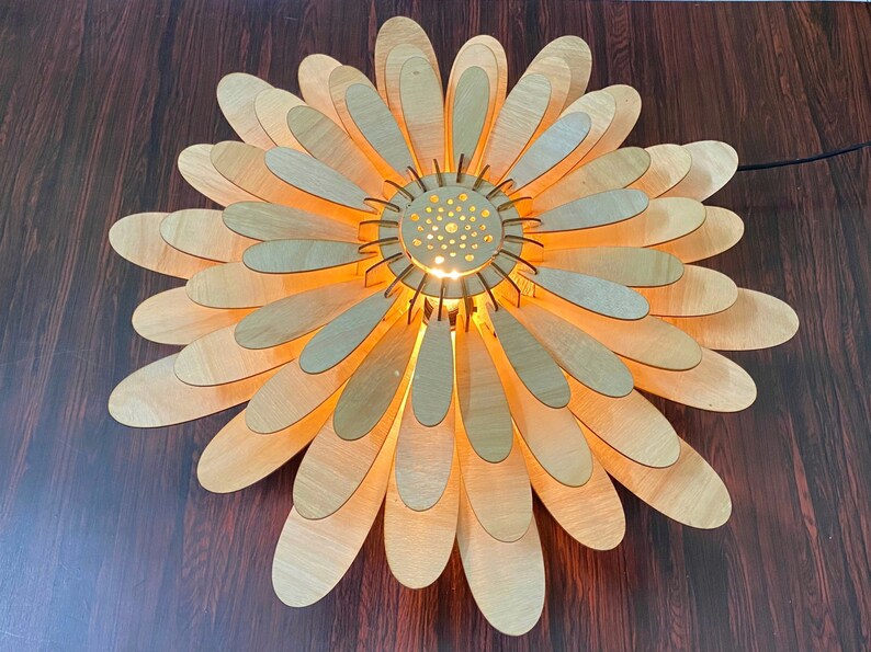 May include: A wooden ceiling light fixture shaped like a daisy with a light bulb in the center. The light fixture is made of many thin, overlapping pieces of wood that are painted a light brown color.