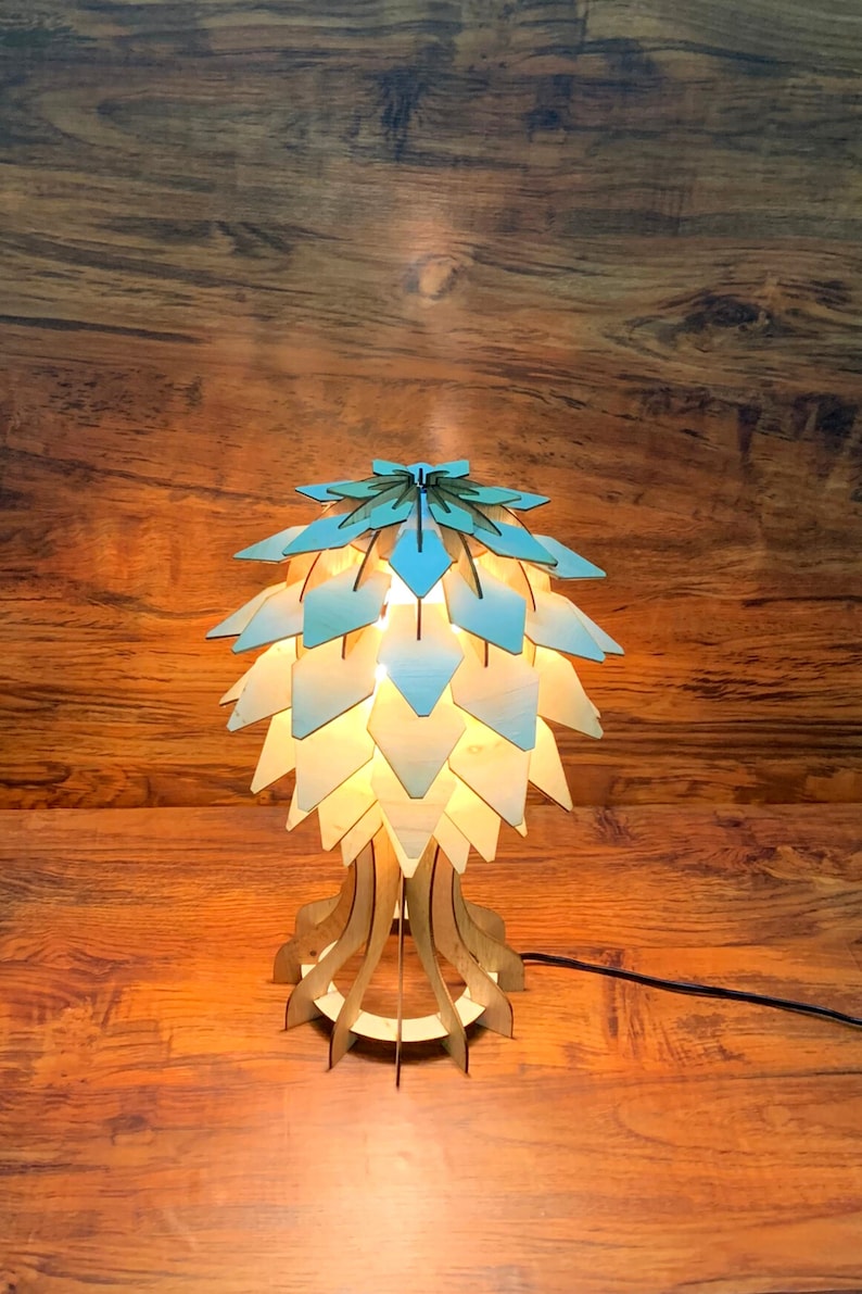 Op de afbeelding: Een houten tafellamp met een geometrische kap in lichtblauw en wit. De lamp heeft een uniek, organisch ontwerp met een voet die lijkt op een boomstam.