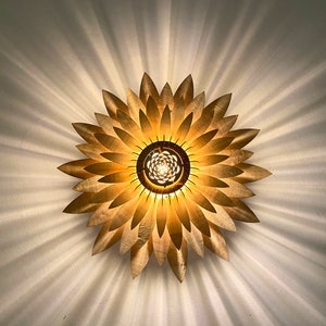 Op de afbeelding: Een gouden metalen plafondlamp in de vorm van een zonnebloem met een centrale gloeilamp.