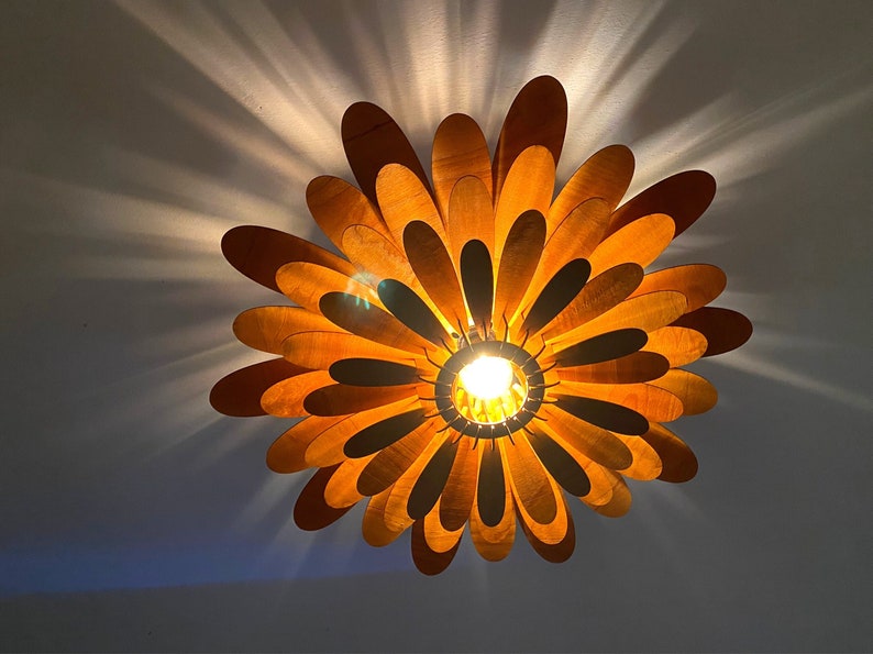 May include: A wooden ceiling light fixture shaped like a flower with multiple layers of petals. The petals are a light brown colour and the light fixture is illuminated by a single light bulb in the centre.