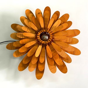 May include: A wooden flower-shaped lamp with a light bulb in the centre. The lamp is made of many thin, overlapping pieces of wood, creating a layered effect. The wood is a light brown colour.