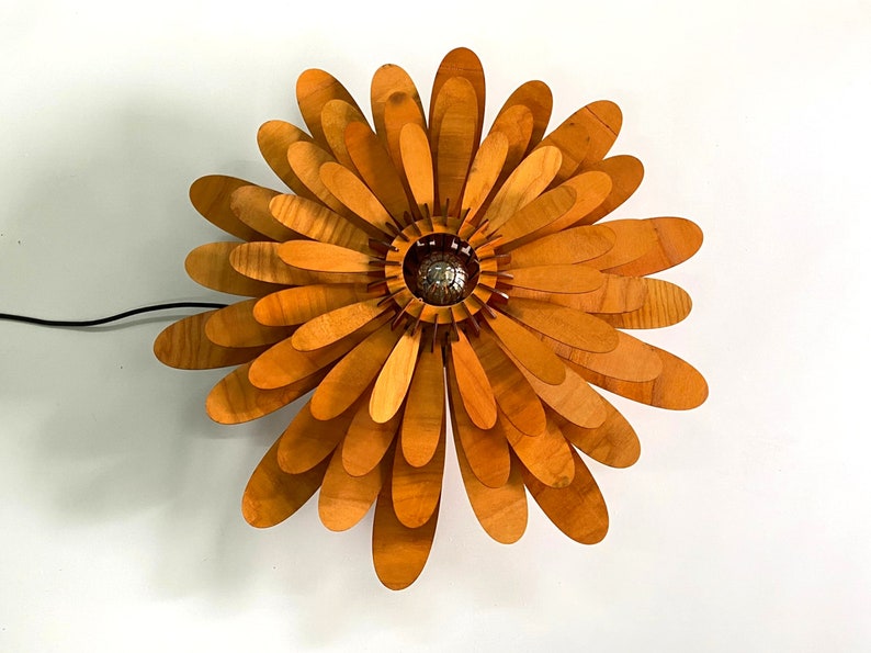 May include: A wooden flower-shaped lamp with a light bulb in the centre. The lamp is made of many thin, overlapping pieces of wood, creating a layered effect. The wood is a light brown colour.