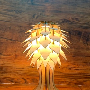 Peut inclure: Une lampe de table en bois au design unique. La lampe pr&eacute;sente un abat-jour en forme de feuilles superpos&eacute;es et un pied d&eacute;taill&eacute;. La lumi&egrave;re chaude cr&eacute;e une ambiance chaleureuse. La lampe est en bois clair.