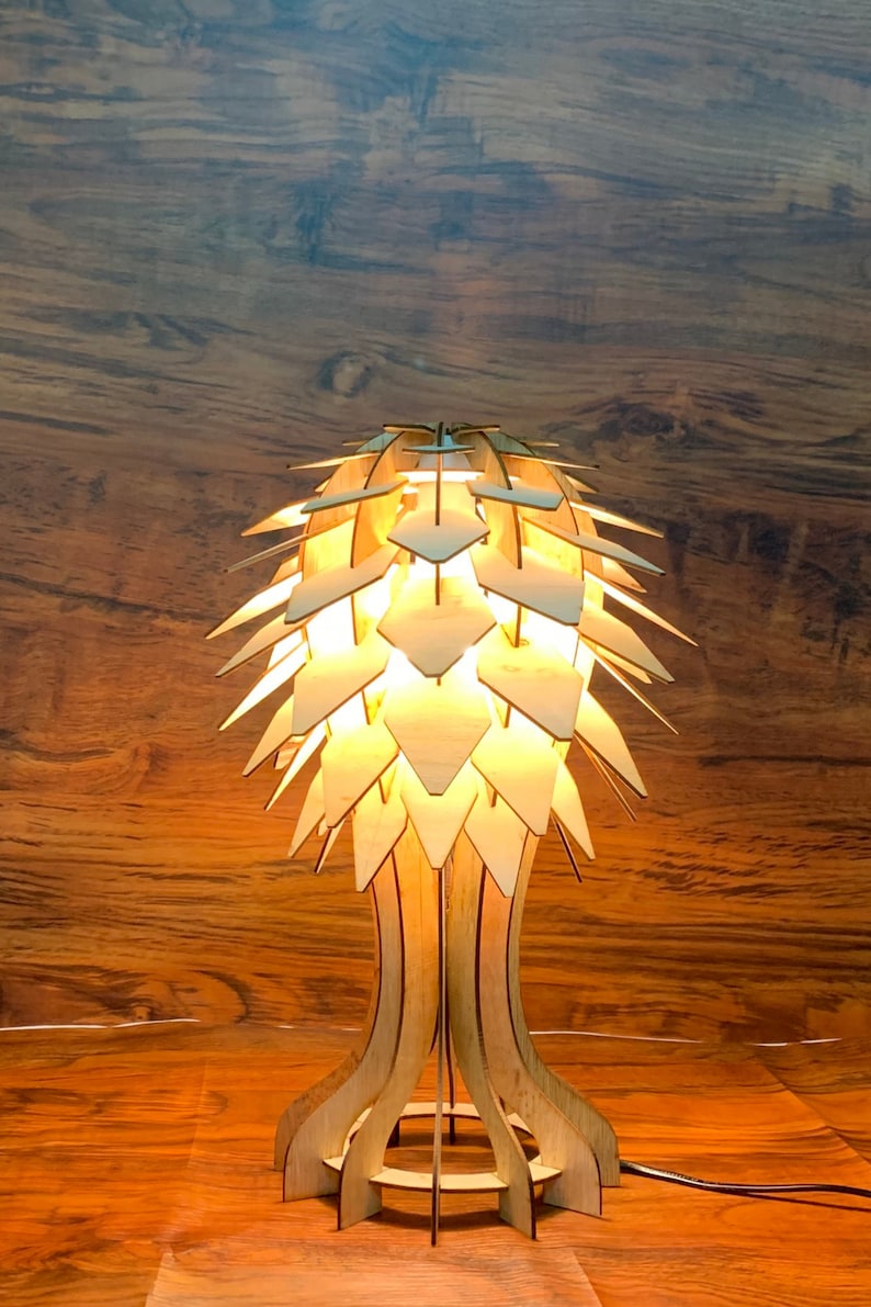 Peut inclure: Une lampe de table en bois au design unique. La lampe pr&eacute;sente un abat-jour en forme de feuilles superpos&eacute;es et un pied d&eacute;taill&eacute;. La lumi&egrave;re chaude cr&eacute;e une ambiance chaleureuse. La lampe est en bois clair.
