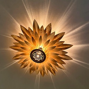 May include: A wooden ceiling light fixture shaped like a sunflower with multiple layers of petals. The light is on and casting a warm glow.