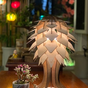 Peut inclure: Lampe de table en bois au design unique, avec un abat-jour g&eacute;om&eacute;trique en couches. La lampe est en bois clair et &eacute;met une lueur chaleureuse. Une petite plante en pot avec des fleurs roses est &agrave; proximit&eacute;. Le texte "ENLIGHTENINGDECOR.COM" est visible.