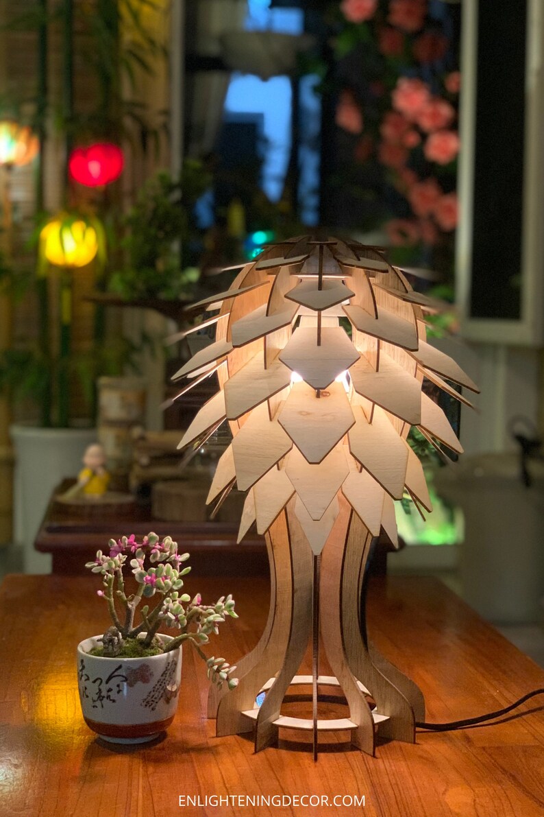 Peut inclure: Lampe de table en bois au design unique, avec un abat-jour g&eacute;om&eacute;trique en couches. La lampe est en bois clair et &eacute;met une lueur chaleureuse. Une petite plante en pot avec des fleurs roses est &agrave; proximit&eacute;. Le texte "ENLIGHTENINGDECOR.COM" est visible.