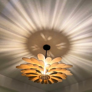 May include: A wooden ceiling light fixture shaped like a flower with light brown petals and a black metal stem. The light casts a pattern of light and shadow on the white ceiling.
