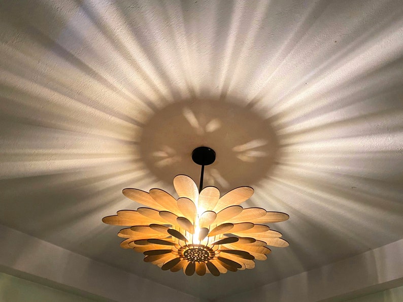 May include: A wooden ceiling light fixture shaped like a flower with light brown petals and a black metal stem. The light casts a pattern of light and shadow on the white ceiling.