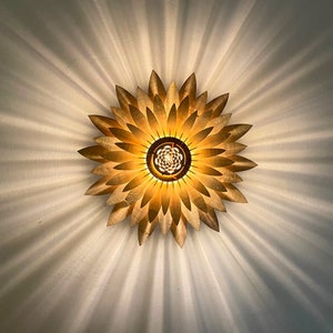 Op de afbeelding: Een gouden plafondlamp in de vorm van een zonnebloem met meerdere bloemblaadjes. De lamp geeft een warm licht op het witte plafond.