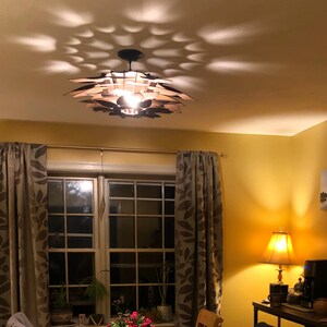 Może przedstawiać: Drewniana lampa sufitowa w kolorze brązowym z unikalnym geometrycznym wzorem rzuca wzorzysty cień na sufit. Lampa jest włączona i oświetla pomieszczenie.