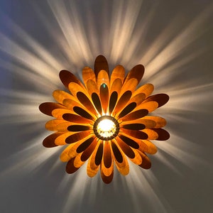 May include: A wooden ceiling light fixture shaped like a flower with multiple layers of petals. The light is on and the petals are a warm brown colour.