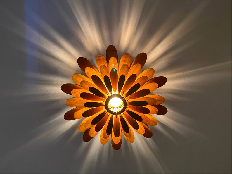 May include: A wooden ceiling light fixture shaped like a flower with multiple layers of petals. The light is on and the petals are a warm brown colour.