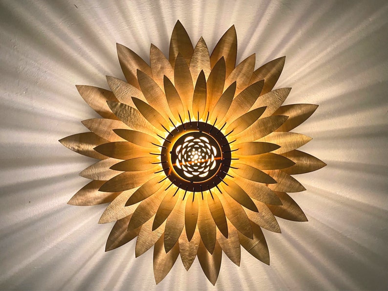 Op de afbeelding: Een gouden plafondlamp in de vorm van een zonnebloem met een ronde lichtbron in het midden.