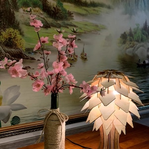 Op de afbeelding: Een houten lamp met een uniek, geometrisch ontwerp dat lijkt op een dennenappel. De lamp is aan en geeft een warm licht. Een vaas met roze bloemen staat op een houten tafel naast de lamp.