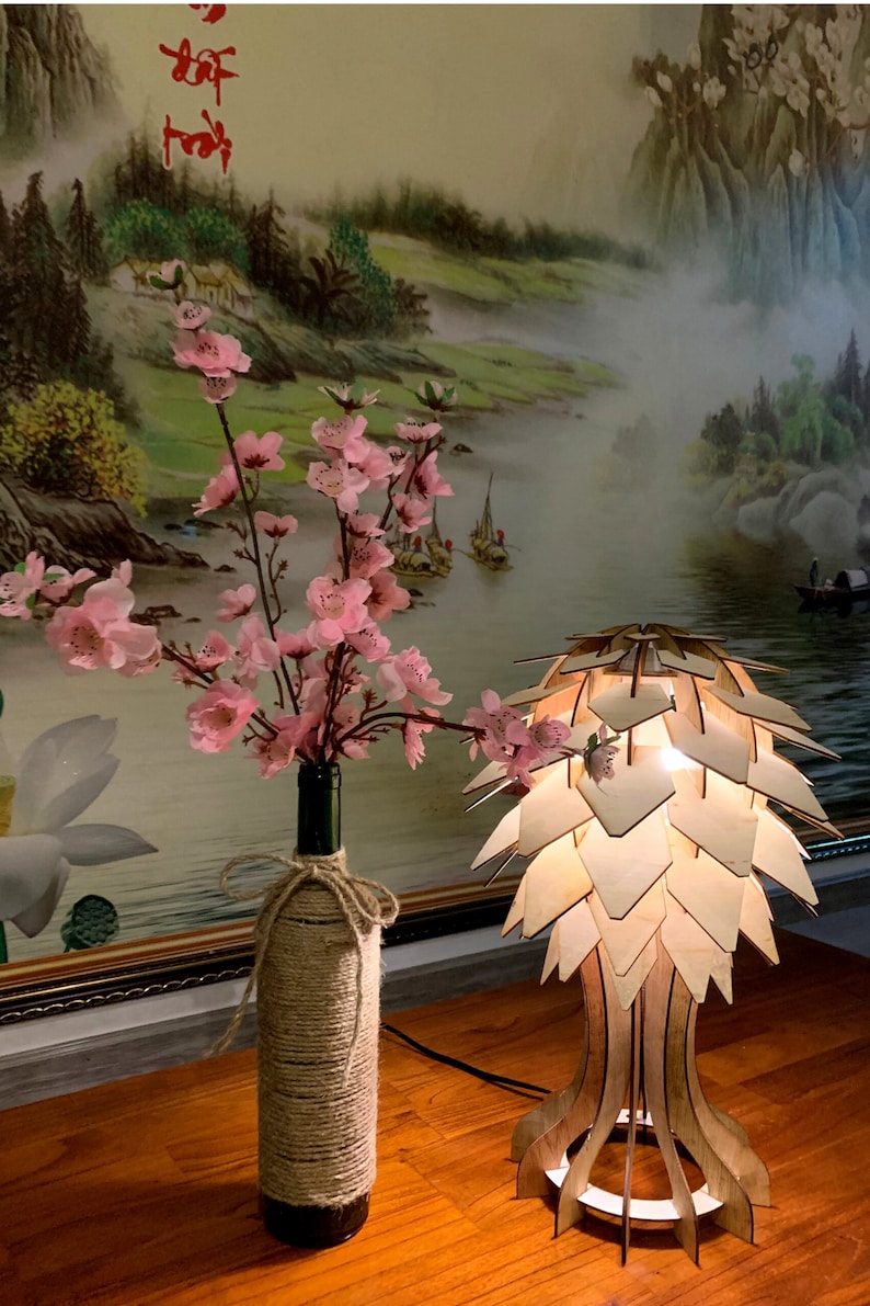 Op de afbeelding: Een houten lamp met een uniek, geometrisch ontwerp dat lijkt op een dennenappel. De lamp is aan en geeft een warm licht. Een vaas met roze bloemen staat op een houten tafel naast de lamp.