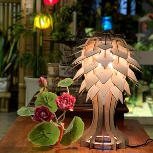 Op de afbeelding: Een houten tafellamp met een uniek ontwerp dat lijkt op een dennenappel. De lamp is gemaakt van licht hout en heeft een warme, uitnodigende gloed. De lamp staat op een houten tafel met een kleine potplant ervoor.