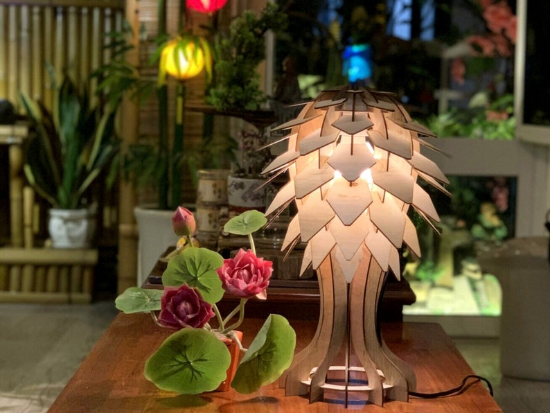 Op de afbeelding: Een houten tafellamp met een uniek ontwerp dat lijkt op een dennenappel. De lamp is gemaakt van licht hout en heeft een warme, uitnodigende gloed. De lamp staat op een houten tafel met een kleine potplant ervoor.