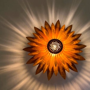Puede incluir: Una l&aacute;mpara de madera con forma de girasol con una luz c&aacute;lida y brillante que emana del centro. La l&aacute;mpara est&aacute; hecha de finos p&eacute;talos de madera superpuestos.