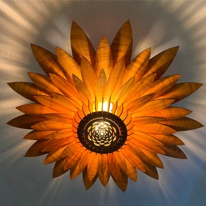 Puede incluir: Una l&aacute;mpara de techo de madera con forma de girasol con un c&aacute;lido brillo amarillo. La l&aacute;mpara est&aacute; hecha de finas piezas de madera superpuestas que se asemejan a los p&eacute;talos de un girasol.