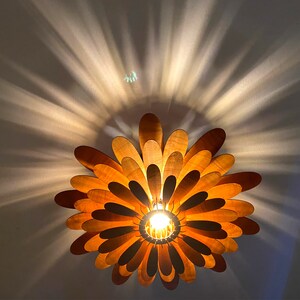 May include: A wooden ceiling light fixture shaped like a flower with multiple layers of petals. The light is on and casts a warm glow.