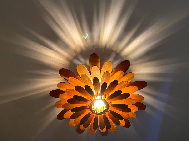 May include: A wooden ceiling light fixture shaped like a flower with multiple layers of petals. The light is on and casts a warm glow.