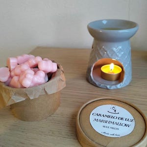 Puede incluir: Un quemador de cera de cerámica gris con una vela encendida, junto a un pequeño recipiente con ceras perfumadas rosas. Un recipiente redondo con una etiqueta que dice "Caramelo de Luz Marshmallow Wax Melts".