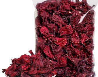 Può includere: Un sacchetto di plastica trasparente pieno di fiori di ibisco rossi secchi. I fiori sono di un rosso intenso e hanno una consistenza leggermente rugosa. Il sacchetto è su una superficie bianca.