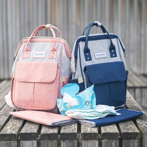 May include: Two diaper bags, one pink and one blue, with the text "Dawson Cooper" on the front. The bags are sitting on a wooden surface with a changing mat and wipes.