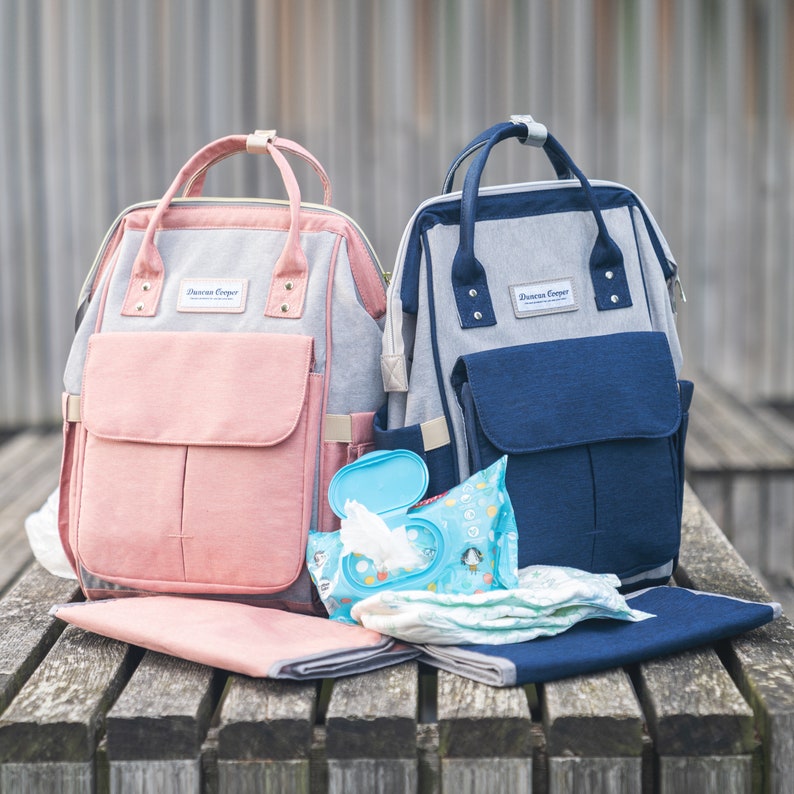 May include: Two diaper bags, one pink and one blue, with the text "Dawson Cooper" on the front. The bags are sitting on a wooden surface with a changing mat and wipes.