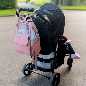 May include: A pink and gray diaper bag with gold accents is attached to a black stroller with a black canopy. The stroller is on a paved road.