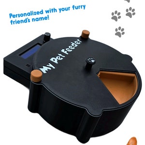 May include: Black automatic pet feeder with a digital display and five separate compartments. The feeder has a brown plastic rim and the text "My Pet Feeder" is printed on the front. Two pets, a cat and a dog, are looking at the feeder.