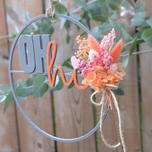 Könnte beinhalten: Ein kreisförmiger grauer Wandbehang mit dem grauen Text OH und dem orangefarbenen Text hi, akzentuiert durch ein kleines rosa Herz und einen Strauß getrockneter Blumen in Orange- und Rosatönen, aufgehängt an einer dünnen Schnur aus Jutegarn.
